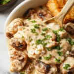 A close-up of a creamy mushroom chicken dish served on a white plate, featuring tender chicken breasts smothered in a rich, velvety mushroom sauce with sautéed mushroom slices and fresh herbs, garnished with chopped parsley.